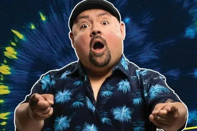 Comic Gabriel Iglesias in a colorful blue and black shirt stands in front of a tie-dye background with a surprised look on his face