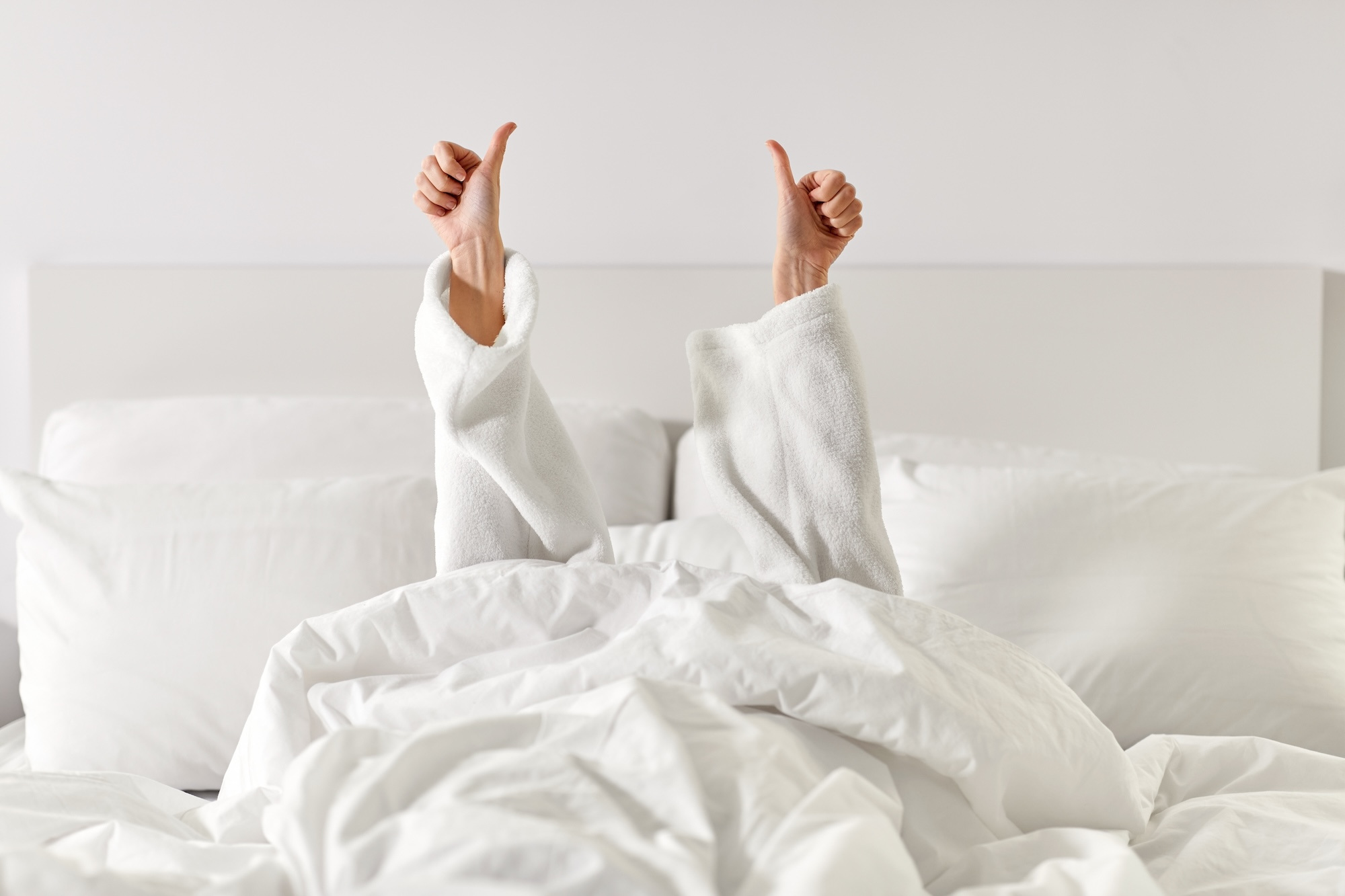 A view from the end of a bed looking up to a person lying in a fluffy white bed, arms stretched toward the ceiling giving a thumbs up with both hands